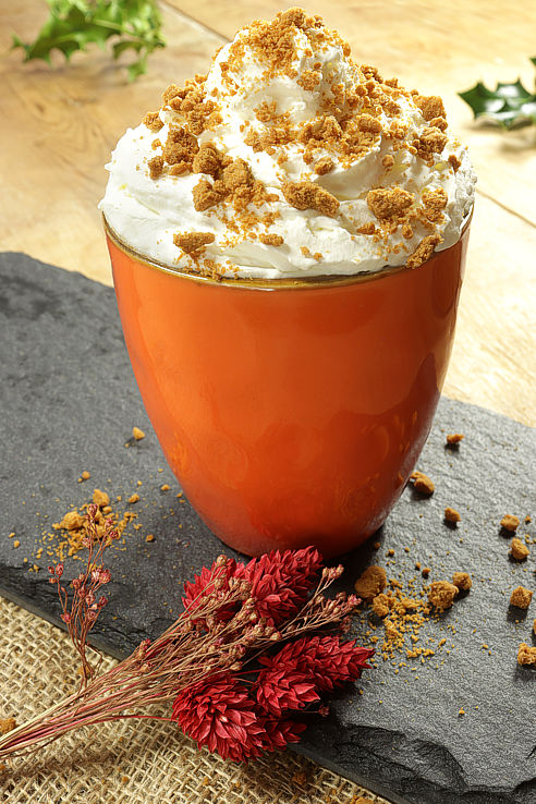 a towering bright orange mug of biscoff latte with a whipped cream topping and biscuit crumbs