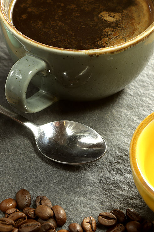 a cup of steaming black coffee with a speen and coffee beans on dark slate