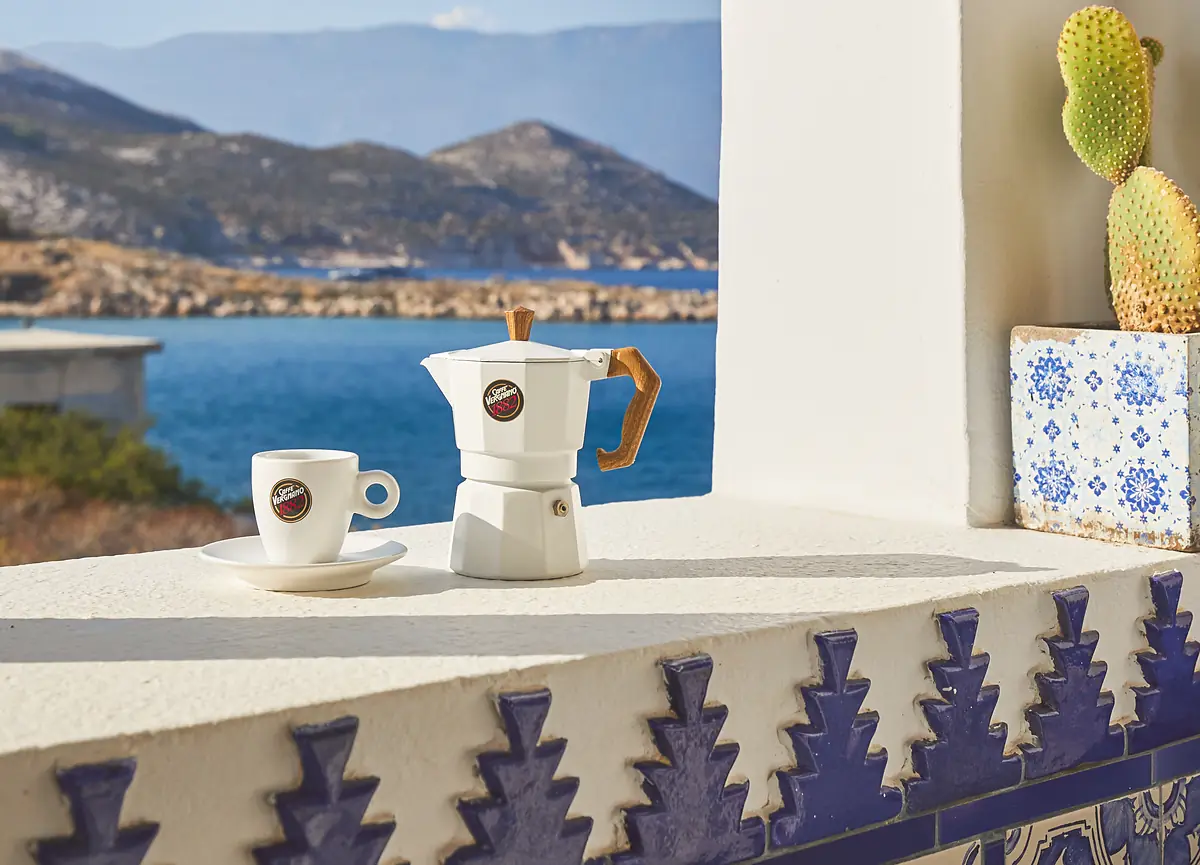 A Caffè Vergnano espresso cup and moka pot on a sunlit terrace above the sea